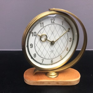 Puede incluir: Un reloj de escritorio dorado con una esfera blanca y números romanos negros. El reloj tiene un patrón de diamante en la esfera y está montado sobre una base de madera.