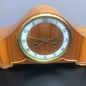 Op de afbeelding: Een houten schoorsteenklok met een witte wijzerplaat en gouden accenten. De klok heeft een gebogen bovenkant en een rechthoekige basis. De wijzerplaat van de klok heeft zwarte Romeinse cijfers als uurmarkeringen en zwarte wijzers. Op de wijzerplaat van de klok staat ook het woord "Hermle".
