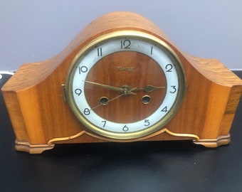 May include: A wooden mantel clock with a white face and gold accents. The clock has a curved top and a rectangular base. The clock face has black Roman numeral hour markers and black hands. The clock face also has the word "Hermle" printed on it.