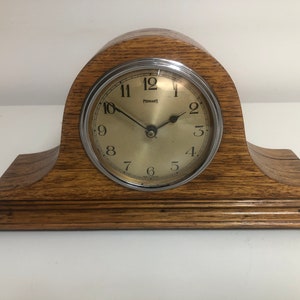 May include: A vintage wooden mantel clock with a light brown finish. The clock face is gold with black numerals and hands, and a silver bezel. The clock face reads "FERRANTI". The clock has a curved top and a rectangular base.