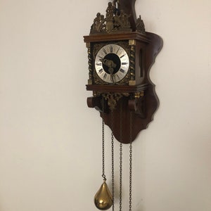 Könnte beinhalten: Eine antike Wanduhr aus Holz mit einem Messingzifferblatt und verzierten Details. Die Uhr hat ein Pendel mit drei Gewichten und ein Zifferblatt mit römischen Ziffern.
