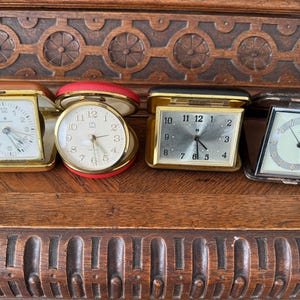May include: Four vintage alarm clocks with different colored cases. The clocks have white faces with black numbers and hands. The clocks are arranged in a row on a wooden surface.