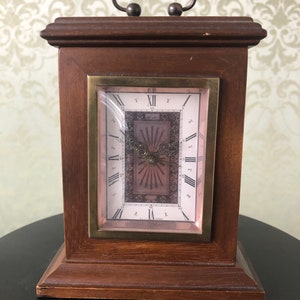 May include: A wooden clock with a brass face and Roman numeral markings. The clock has a decorative design on the face and a handle on the top.