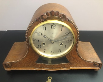 May include: A wooden mantel clock with a brass face and black hands. The clock has a Roman numeral clock face with a white background. The clock is sitting on a wooden base.