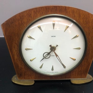 Peut inclure: Une horloge Smiths vintage de 8 jours avec un cadran blanc et des aiguilles dorées. L'horloge est logée dans un boîtier en bois avec des pieds en laiton. Le cadran de l'horloge indique "Smiths 8-day Made in England".