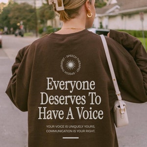 Puede incluir: Una sudadera marr&oacute;n con el texto "Everyone Deserves To Have A Voice" y las palabras "Speech Therapist" en un c&iacute;rculo alrededor de un gr&aacute;fico de sol. El texto debajo del mensaje principal dice: "Your voice is uniquely yours, communication is your right."