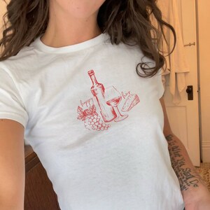 May include: White t-shirt with a red graphic of a wine bottle, glass of wine, grapes, and cheese. The text "happy hour" is written on the shirt.