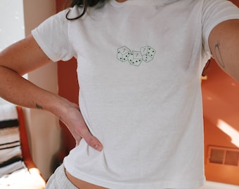 Puede incluir: Camiseta blanca con un diseño bordado verde de tres dados con el número siete en cada cara.
