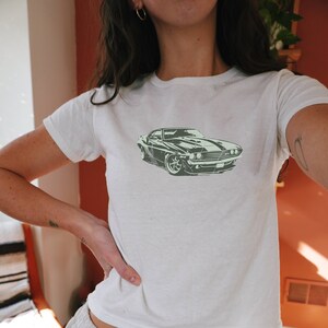 May include: White crop top with a green car graphic. The t-shirt is paired with light gray sweatpants. The car graphic is a vintage muscle car.