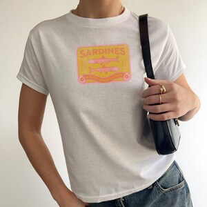 May include: White t-shirt with a graphic of a sardine can label that says "Sardines in Extra Virgin Olive Oil".