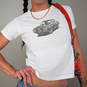 May include: White t-shirt with a black and green muscle car graphic. The car has a black hood and a white stripe down the side.