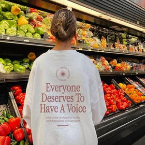 Puede incluir: Sudadera blanca de manga larga con el texto "Everyone Deserves To Have A Voice" en letras rosas. El texto est&aacute; rodeado de un c&iacute;rculo con un gr&aacute;fico de sol. El texto debajo del c&iacute;rculo dice "Your voice is uniquely yours, communication is your right."