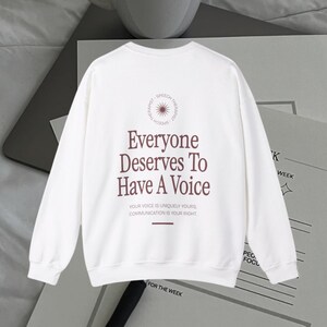 Puede incluir: Sudadera blanca de manga larga con un estampado gr&aacute;fico marr&oacute;n que dice "Everyone Deserves To Have A Voice." El gr&aacute;fico tambi&eacute;n incluye un c&iacute;rculo con un dise&ntilde;o de sol radiante y las palabras "Speech Therapist."