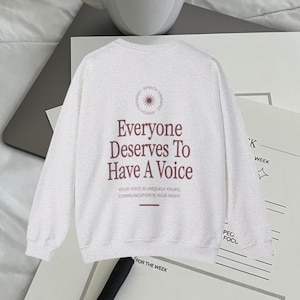 Puede incluir: Una sudadera gris claro de manga larga y cuello redondo. La parte posterior de la sudadera tiene un estampado gr&aacute;fico que dice "Everyone Deserves To Have A Voice." El gr&aacute;fico tambi&eacute;n incluye un c&iacute;rculo con un dise&ntilde;o de sol radiante y las palabras "Speech Therapist" escritas alrededor del c&iacute;rculo. El texto debajo del gr&aacute;fico dice "Your voice is uniquely yours. Communication is your right."
