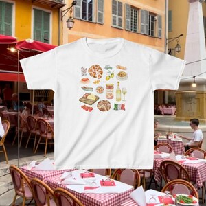 May include: White t-shirt with a watercolor illustration of Italian food, including pizza, bread, pasta, tomatoes, olives, cheese, wine, and a jar of tomato sauce. The illustration is surrounded by a white border.