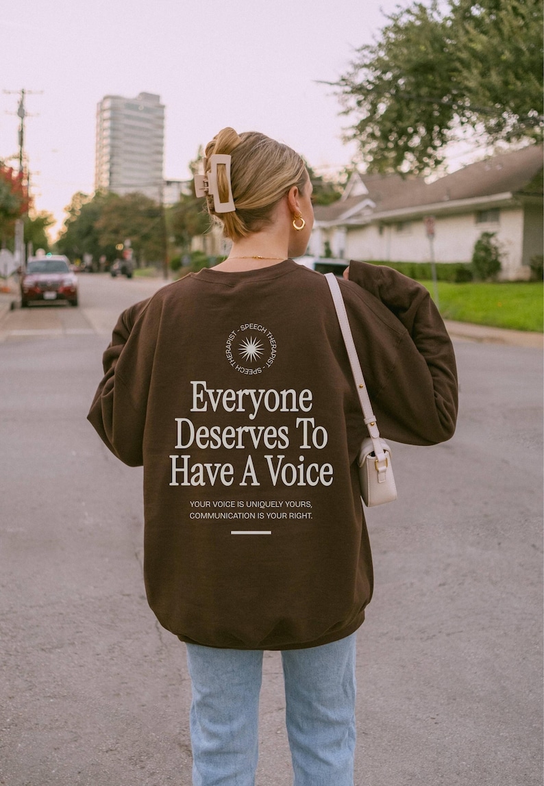 Puede incluir: Una sudadera marr&oacute;n con el texto "Everyone Deserves To Have A Voice" y las palabras "Speech Therapist" en un c&iacute;rculo alrededor de un gr&aacute;fico de sol. El texto debajo del mensaje principal dice: "Your voice is uniquely yours, communication is your right."