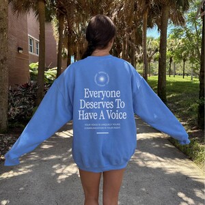 Puede incluir: Una sudadera azul claro con el texto "Everyone Deserves To Have A Voice" y el texto "Your voice is uniquely yours. Communication is your right." impreso en la parte posterior.