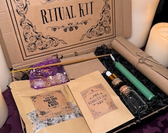 May include: A brown cardboard box with the words "The Moonchild Tarot UK" and "Ritual Kit" printed on the front. The box is open and contains a variety of items, including a small bag of bath salts, a small bottle of oil, a small bottle of dried herbs, a green candle, and a wooden stick.