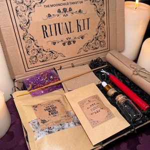 Puede incluir: Una caja de cartón con el texto "THE MOONCHILD TAROT UK" y "RITUAL KIT" impreso en la tapa. La caja contiene una variedad de artículos, incluyendo una pequeña bolsa de sales de baño, una vela, una pequeña botella de aceite esencial, una pequeña bolsa de hierba gatera y un rollo.