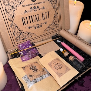 以下が含まれることがあります： 蓋に「THE MOONCHILD TAROT UK」と「RITUAL KIT」と書かれた茶色の段ボール箱。箱が開いており、バスソルトの小さな袋、バラの花びらの袋、エッセンシャルオイルの瓶、ピンクのキャンドル、木の棒、紫と銀のデザインの小さなポーチなど、さまざまなアイテムが入っています。