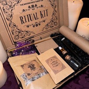 Puede incluir: Una caja de cartón marrón con las palabras "THE MOONCHILD TAROT UK" y "RITUAL KIT" impresas en la tapa. La caja está abierta y contiene varios artículos, incluyendo una pequeña bolsa de sales de baño, una botella de aceite esencial, un pequeño frasco de cristales y un trozo de papel enrollado. La caja está rodeada de velas blancas.