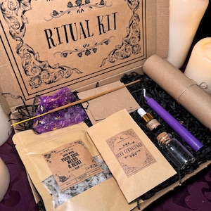 Puede incluir: Una caja de cartón marrón con las palabras "The Moonchild Tarot UK" y "Ritual Kit" impresas en la tapa. La caja está abierta y contiene una variedad de artículos, incluyendo una pequeña bolsa de sales de baño, una vela morada, una pequeña botella de aceite esencial, una pequeña bolsa de hierbas y una pequeña bolsa de cristales. La caja está sobre una superficie morada.