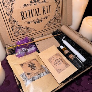 Puede incluir: Una caja de cartón marrón con las palabras "THE MOONCHILD TAROT UK" y "RITUAL KIT" impresas en la parte superior. La caja está abierta y contiene una variedad de artículos, incluyendo una pequeña bolsa de sales de baño, una vela, una botella de aceite, una pequeña bolsa de hierbas y un trozo de papel con las palabras "MOTHERWORT" impresas en él.