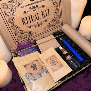 Puede incluir: Una caja de cartón marrón con un diseño floral en blanco y negro y el texto "The Moonchild Tarot UK Ritual Kit". La caja está abierta y contiene varios artículos, incluyendo una pequeña bolsa de sales de baño, una botella de aceite esencial, una vela azul, una pequeña bolsa de hierbas y un rollo de papel pergamino.