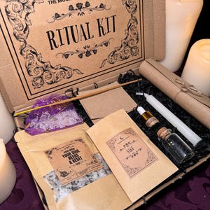 以下が含まれることがあります： 「The Moonchild Tarot UK Ritual Kit」と印刷された茶色の段ボール箱。箱が開いており、小さなバスソルトの袋、小さなセージの瓶、白いろうそく、木製の棒など、さまざまなアイテムが入っています。箱は紫色の表面の上に置かれています。