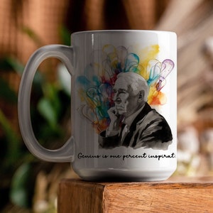 May include: White ceramic mug featuring a black and white portrait of a man with colorful abstract art. The mug has the quote "Genius is one percent inspiration". The mug is on a wooden surface.