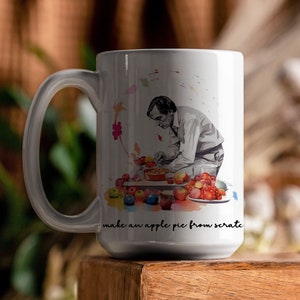 May include: White ceramic mug featuring a black and white illustration of a chef preparing apples for a pie. The mug has the text "make an apple pie from scratch" and colorful watercolor accents.