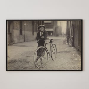 May include: A black and white photograph of a young boy standing with his bicycle in a city alleyway. The boy is wearing a cap and a jacket. The bicycle has a front wheel with a white tire and a back wheel with a black tire.