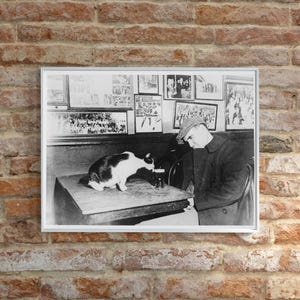 May include: Black and white photograph of a man in a coat and hat sitting at a table with a black and white cat. The man is looking at the cat, which is drinking from a glass. The photo is framed and hanging on a brick wall.