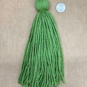 May include: A green yarn tassel with a round top. The tassel is about 4 inches long and is shown next to a US quarter for scale.