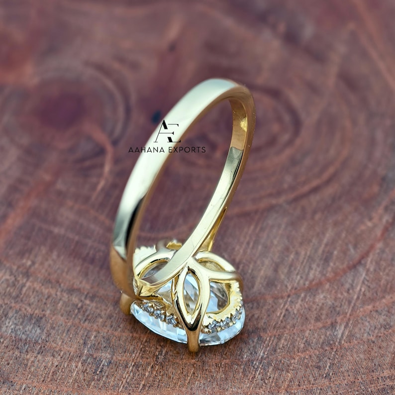 May include: A gold-colored ring with a large, clear, oval-shaped gemstone. The ring has a detailed setting with smaller, clear stones around the main gem. The band is smooth and polished. The text "AAHANA EXPORTS" is visible on the ring.