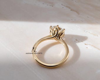 May include: A gold engagement ring with a large, clear gemstone. The ring has a delicate, floral-inspired setting that holds the stone securely. The band is smooth and polished, reflecting the light. The ring is set against a marble background.