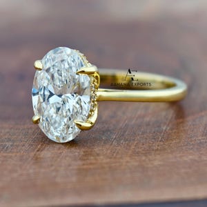 May include: A gold engagement ring with a large, oval-cut diamond as the centerpiece. The diamond is clear and faceted, set in a gold band with smaller accent stones. The ring is displayed on a wooden surface, showcasing its sparkle and design.
