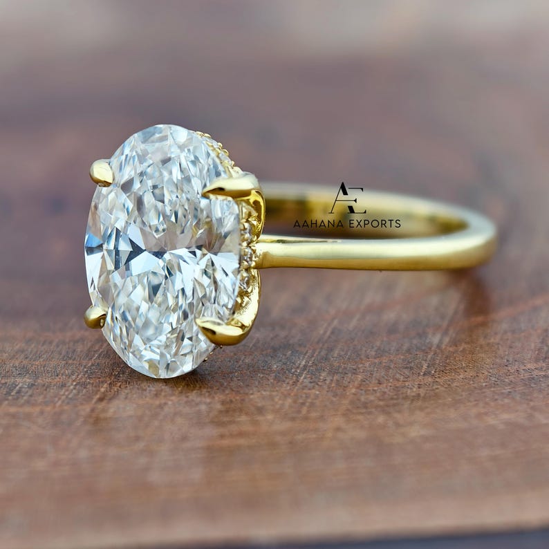 May include: A gold engagement ring with a large, oval-cut diamond as the centerpiece. The diamond is clear and faceted, set in a gold band with smaller accent stones. The ring is displayed on a wooden surface, showcasing its sparkle and design.