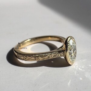 May include: A gold ring with an oval-shaped diamond. The ring has an engraved floral pattern on the band. The diamond is set in a bezel setting. The ring is shown on a white surface.