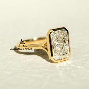 May include: A gold ring with a large, rectangular-cut diamond. The diamond is set in a bezel setting, and the band is polished gold. The ring is displayed on a light surface. The text "AAHANA EXPORTS" is visible on the ring.