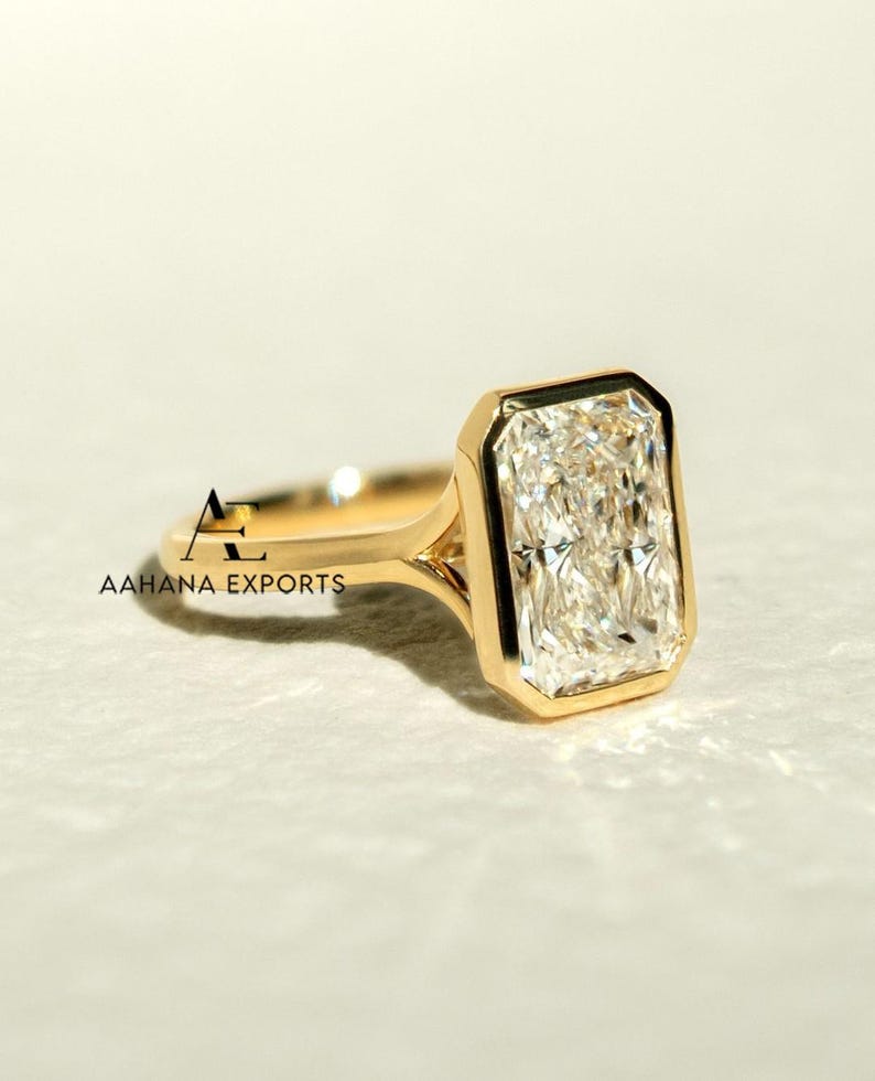 May include: A gold ring with a large, rectangular-cut diamond. The diamond is set in a bezel setting, and the band is polished gold. The ring is displayed on a light surface. The text "AAHANA EXPORTS" is visible on the ring.