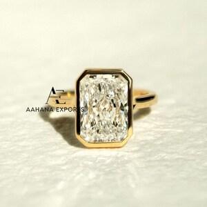 May include: A gold ring with a large, rectangular, clear gemstone. The gemstone is set in a gold bezel. The ring has a simple design. The text "AAHANA EXPORTS" is visible in the lower left corner.