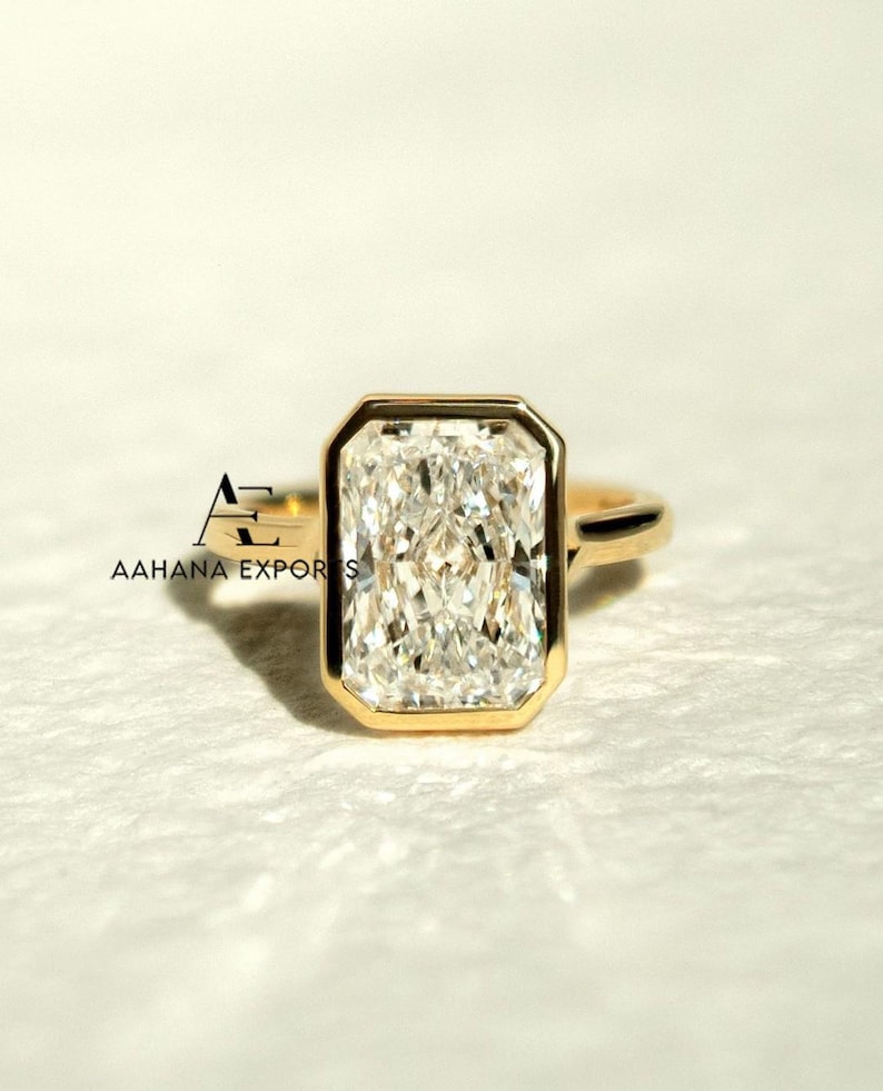May include: A gold ring with a large, rectangular, clear gemstone. The gemstone is set in a gold bezel. The ring has a simple design. The text "AAHANA EXPORTS" is visible in the lower left corner.