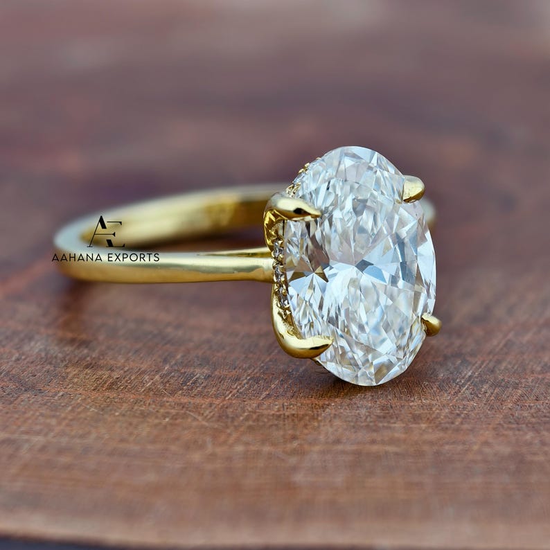 May include: A gold engagement ring with a large, oval-cut diamond. The ring has a delicate band and claw prongs holding the diamond. The brand name "AAHANA EXPORTS" is visible on the ring's band.