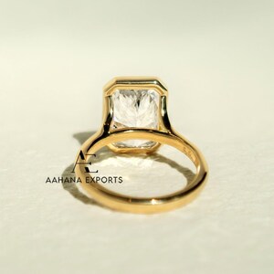 May include: A gold ring featuring a large, rectangular, clear gemstone. The ring has a simple, elegant design with a smooth, polished band. The gemstone is set in a bezel setting. The ring is viewed from the back.