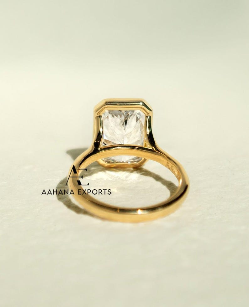 May include: A gold ring featuring a large, rectangular, clear gemstone. The ring has a simple, elegant design with a smooth, polished band. The gemstone is set in a bezel setting. The ring is viewed from the back.