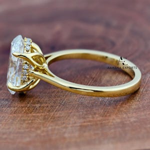 May include: A close-up of a gold ring with a large, clear, oval-cut gemstone. The ring has a delicate band and is set with smaller accent stones around the main stone. The ring is on a wooden surface. The text "AAHANA EXPORTS" is visible.