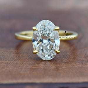 May include: A gold ring featuring a large, oval-cut diamond. The diamond is clear and multifaceted, set with four gold prongs. The ring band is a simple, polished gold. The text "AAHANA EXPORTS" is visible on the ring.