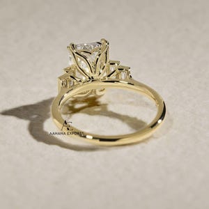 May include: A gold ring with a large, square-cut clear gemstone as the centerpiece, flanked by smaller rectangular stones. The ring's band is a simple, polished gold. The ring is set against a neutral background.