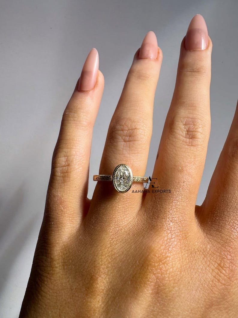 May include: A gold ring with an oval-shaped diamond center stone. The ring is set on a hand with long, polished nails. The band has a delicate design. The ring is from AAHAA EXPORTS.